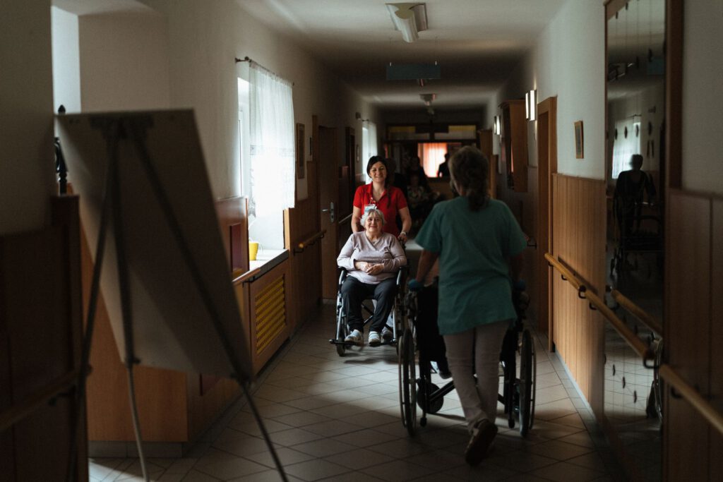 Pflegekraft begleitet eine ältere Frau im Rollstuhl durch den Flur eines Seniorenheims